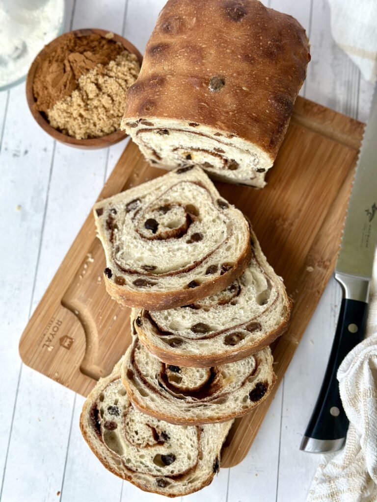 Overhead: Loaf of cinnamon raisin sourdough bread on a wooden cutting board. Several slices sit in front of the loaf.